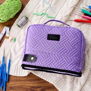 May include: A rectangular purple quilted storage case with a top handle and a black trim. The front features a black rectangular patch with the name Claire embroidered in script. A black tab with a gold-tone snap button secures the front flap.
