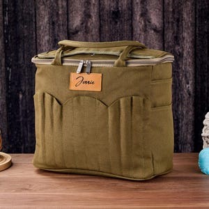 May include: An olive green craft bag with multiple pockets and a top handle. The bag has a zip closure and a leather tag that says "Jennie". The bag is on a wooden surface with yarn and knitting needles.