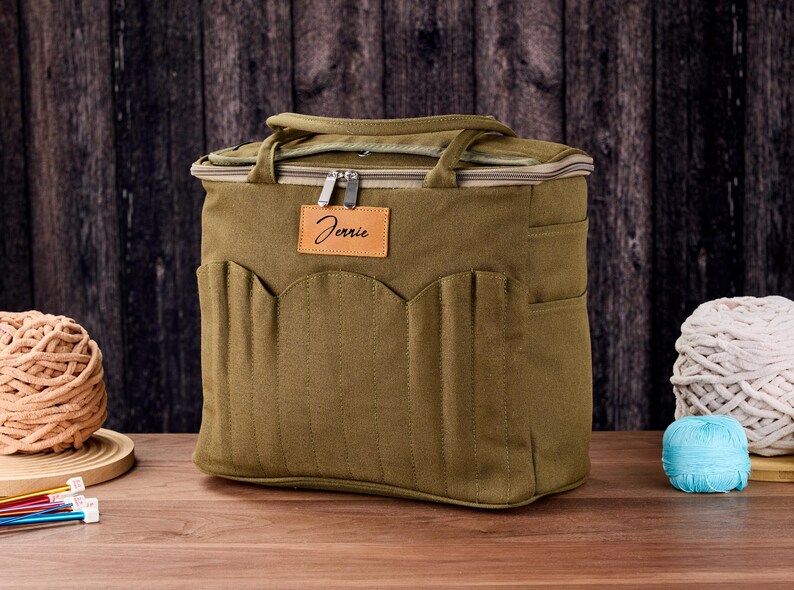 May include: An olive green craft bag with multiple pockets and a top handle. The bag has a zip closure and a leather tag that says "Jennie". The bag is on a wooden surface with yarn and knitting needles.