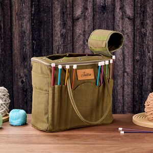 May include: An olive green knitting bag with multiple pockets and a handle. The bag contains colourful knitting needles and has a brown leather patch that says "Justin". Several balls of yarn in various colours are next to the bag.