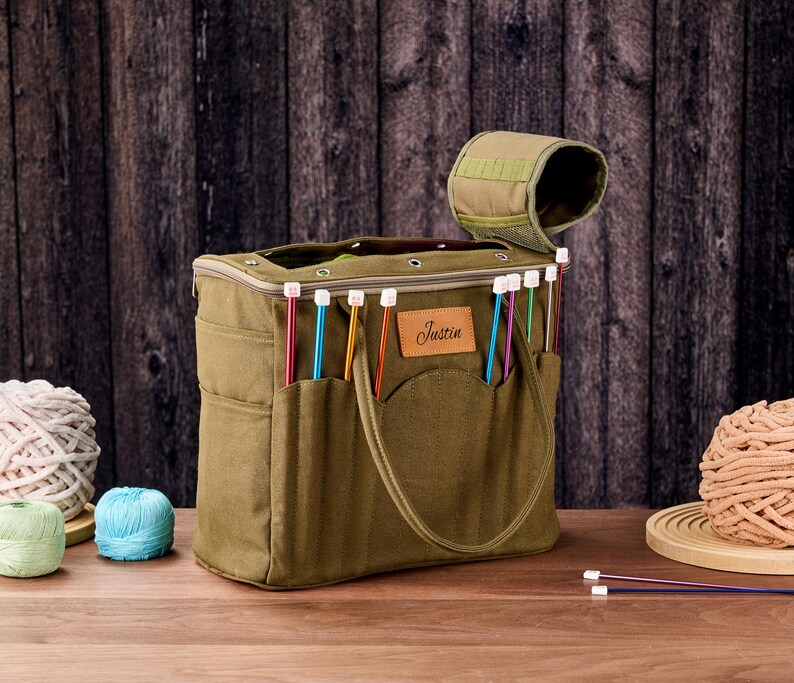 May include: An olive green knitting bag with multiple pockets and a handle. The bag contains colourful knitting needles and has a brown leather patch that says "Justin". Several balls of yarn in various colours are next to the bag.