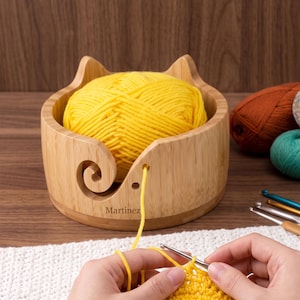 May include: A light-colored wooden yarn bowl with cat-ear shaped edges and a spiral cutout, containing a large ball of bright yellow yarn. The word Martinez is printed on the front. A strand of yellow yarn feeds through the cutout to a crochet hook.