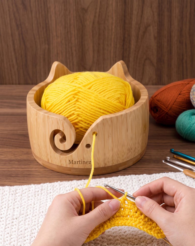 May include: A light-colored wooden yarn bowl with cat-ear shaped edges and a spiral cutout, containing a large ball of bright yellow yarn. The word Martinez is printed on the front. A strand of yellow yarn feeds through the cutout to a crochet hook.
