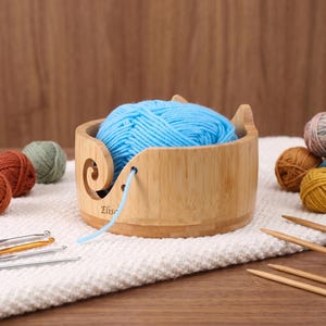 May include: A round bamboo yarn bowl with a spiral cutout and a small hole for feeding yarn, containing a ball of bright blue yarn. The bowl features the name Elise engraved on the side. Several metal crochet hooks and wooden knitting needles are nearby.