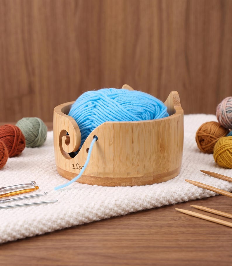 May include: A round bamboo yarn bowl with a spiral cutout and a small hole for feeding yarn, containing a ball of bright blue yarn. The bowl features the name Elise engraved on the side. Several metal crochet hooks and wooden knitting needles are nearby.