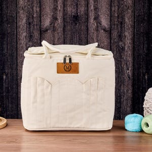May include: Cream-coloured craft bag with a top handle and a front pocket design. The bag has a brown leather tag with the letter "M" on it. Two balls of yarn and two skeins of yarn are visible in the background.