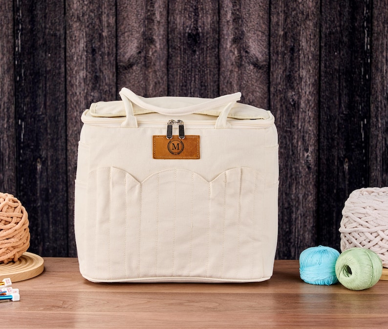 May include: Cream-coloured craft bag with a top handle and a front pocket design. The bag has a brown leather tag with the letter "M" on it. Two balls of yarn and two skeins of yarn are visible in the background.