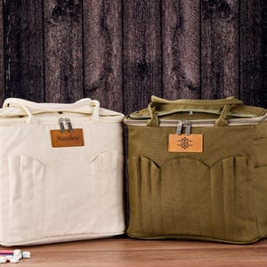 May include: Two canvas tote bags, cream and olive green, with top handles and front pockets. Each bag has a leather patch with text or a design. The cream bag has the name "Matthew" on it.