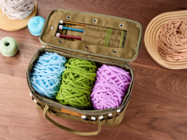 May include: An open olive green craft bag filled with blue, green, and purple yarn. Knitting needles are stored in the lid. Additional yarn balls in white, light blue, and green are on the wooden surface.