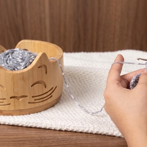 May include: A light brown bamboo yarn bowl featuring a carved cat face design with pointed ears, whiskers, and a smiling mouth. The bowl holds a ball of grey speckled yarn, which is being used for a crochet project with a metal crochet hook.
