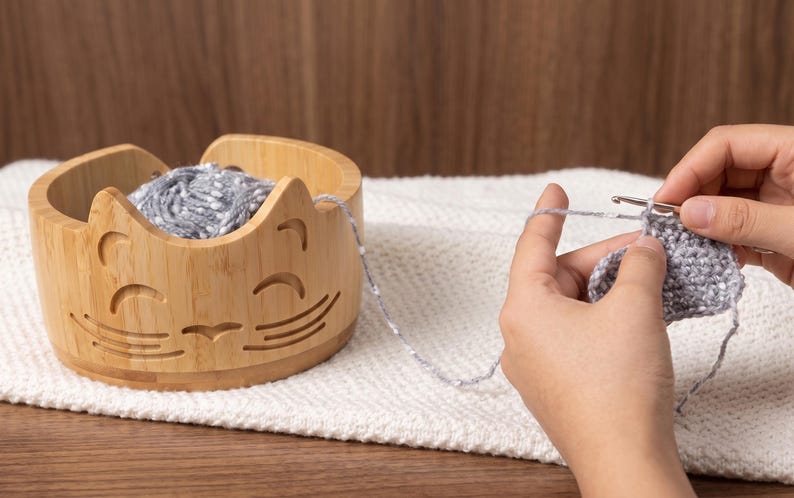 May include: A light brown bamboo yarn bowl featuring a carved cat face design with pointed ears, whiskers, and a smiling mouth. The bowl holds a ball of grey speckled yarn, which is being used for a crochet project with a metal crochet hook.
