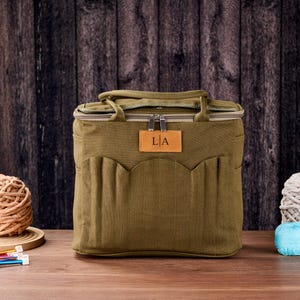 May include: An olive green tote bag with a brown leather tag displaying the initials "L|A". The bag has a zip closure, carrying handles, and multiple exterior pockets. Knitting supplies, including yarn and needles, are visible.