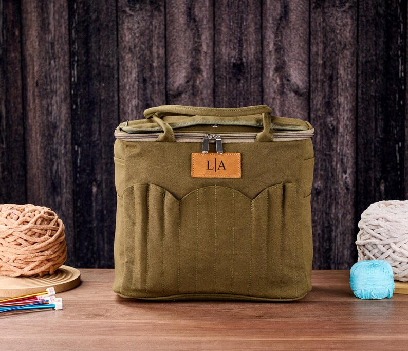 May include: An olive green tote bag with a brown leather tag displaying the initials "L|A". The bag has a zip closure, carrying handles, and multiple exterior pockets. Knitting supplies, including yarn and needles, are visible.