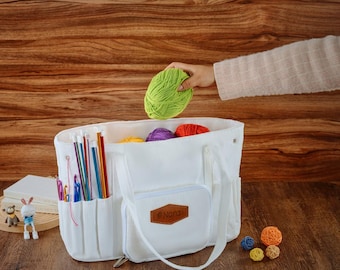 May include: White canvas tote bag with multiple pockets and a brown leather patch that says "Nora". The bag is filled with colorful yarn balls and knitting needles.