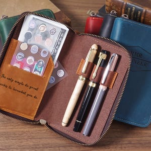 May include: A brown leather zippered organizer case featuring three pen loops, a pocket containing a sheet of small circular stickers, and the engraved text: The only way to do great work is to love what you do. The case is open, revealing its interior.