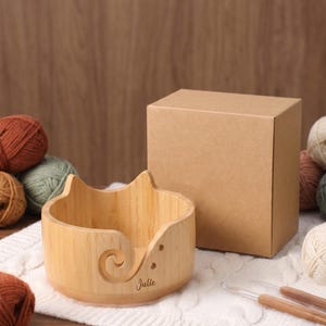May include: A light-colored wooden yarn bowl with cat-ear shaped edges and a spiral cutout. The bowl features the name Julie engraved on the front. It is accompanied by a plain brown cardboard box and two wooden crochet hooks on a white fabric.