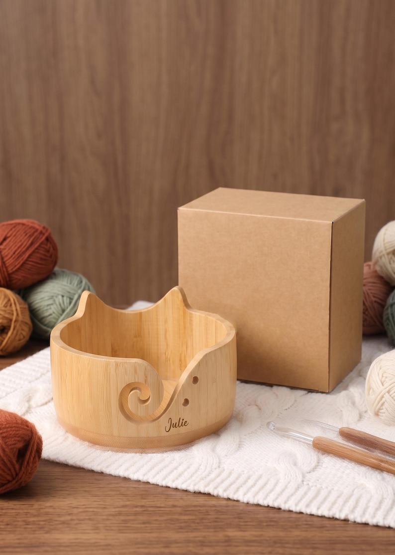 May include: A light-colored wooden yarn bowl with cat-ear shaped edges and a spiral cutout. The bowl features the name Julie engraved on the front. It is accompanied by a plain brown cardboard box and two wooden crochet hooks on a white fabric.