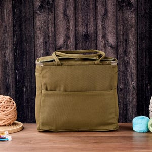 May include: An olive green tote bag with a top handle and front pocket, beside several balls of yarn in various colours. Knitting needles are visible in the foreground. The background is a dark wooden wall.