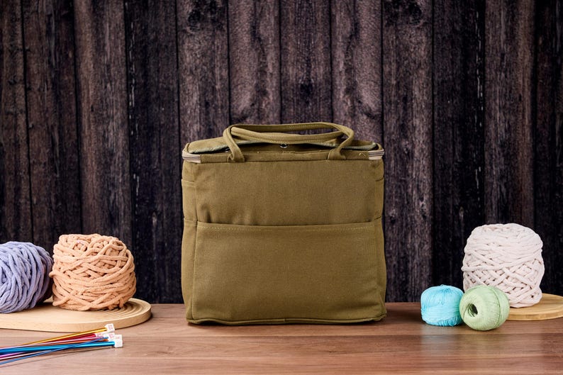 May include: An olive green tote bag with a top handle and front pocket, beside several balls of yarn in various colours. Knitting needles are visible in the foreground. The background is a dark wooden wall.