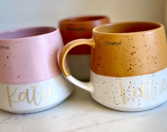 May include: Three ceramic mugs with speckled glaze in pink, orange, and terracotta. Each mug has a white band with the name "Katie" written in gold lettering.