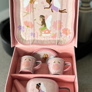 May include: A pink metal suitcase playset with a fairy theme. The set includes a teapot, two teacups, two saucers, and a watering can. The suitcase has a handle and a latch.