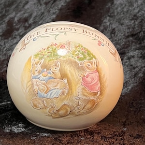 May include: A round, cream-colored ceramic ornament featuring a scene from "The Flopsy Bunnies." The illustration depicts several bunnies in a grassy setting, with the title of the story arching across the top. The ornament has a glossy finish.