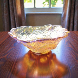 May include: An iridescent, amber-coloured glass bowl with a leaf design. The bowl has a ruffled edge and sits on a dark wooden surface. The bowl's surface reflects light, creating a shimmering effect.