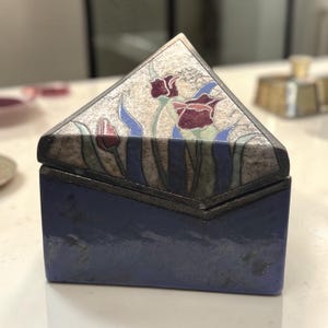 May include: A rectangular box with a solid blue base and a triangular lid. The lid features a textured, light-colored surface decorated with a floral pattern of dark red tulips, green stems, and blue leaves, all outlined in dark metal framing.