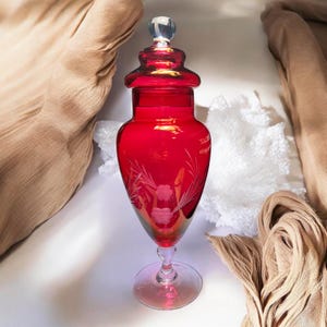 Puede incluir: Un tarro de farmacia de vidrio rojo rubí con un tapón de vidrio transparente. El tarro tiene un diseño floral grabado decorativo y una base de pedestal. El tarro mide aproximadamente 25 cm de altura.