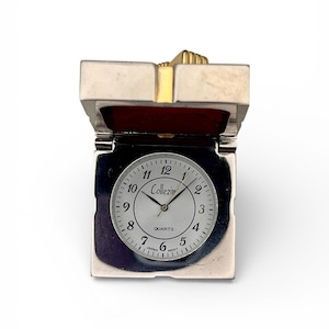 May include: A silver-toned quartz clock with a white face and black numerals. The clock is in a rectangular case with a hinged top. The clock face reads "Collezie" and "Quartz". The top has a gold-colored accent.