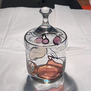 Pode incluir: Um frasco de vidro transparente com tampa, com desenhos de frutas pintados à mão em tons de rosa, laranja e dourado. O frasco tem uma pega transparente na tampa e foi concebido para guardar pequenos objetos. O frasco está sobre uma superfície branca.