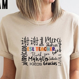 May include: A beige t-shirt with the words "ESL TEACHER" in colorful letters surrounded by words of thanks in different languages, including "Merci", "Shukran", "Gracias", "Thank you", "Mahalo", "Tadhunter", "Tak Kiitos", "Danke", "Obrigado", "Besharisto", and "VIVAH".