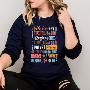 May include: A navy blue long-sleeved sweatshirt with a graphic print of the word "Hello" in multiple languages. The text is in a variety of colors, including pink, blue, and yellow.