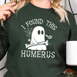 May include: Dark green sweatshirt with the text "I FOUND THIS HUMERUS" and a white ghost graphic holding a bone. The design includes a spider web, spider, and bat. A white mug is held in the hand.