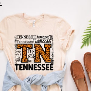 May include: A peach-colored t-shirt with a graphic design featuring the letters "TN" in orange with a leopard print pattern. The letters are surrounded by the word "Tennessee" repeated in various sizes and orientations. The shirt is laid flat on a white surface with a pair of brown leather loafers and a green plant.