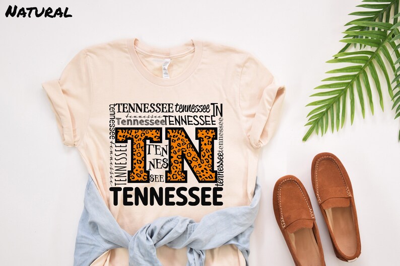 May include: A peach-colored t-shirt with a graphic design featuring the letters "TN" in orange with a leopard print pattern. The letters are surrounded by the word "Tennessee" repeated in various sizes and orientations. The shirt is laid flat on a white surface with a pair of brown leather loafers and a green plant.