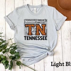 May include: Light blue t-shirt with a graphic design featuring the letters "TN" in orange with a leopard print pattern surrounded by the word "Tennessee" repeated in various sizes and orientations.