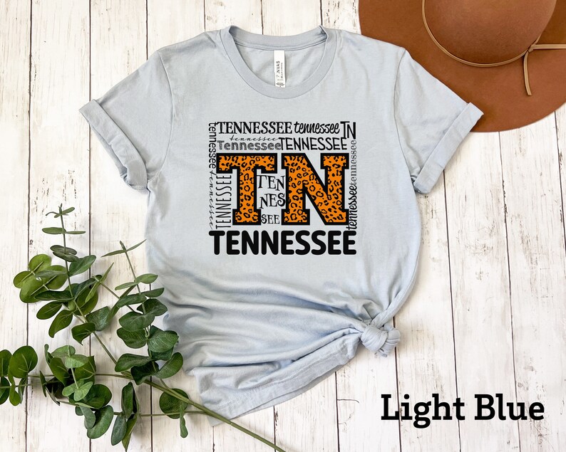 May include: Light blue t-shirt with a graphic design featuring the letters "TN" in orange with a leopard print pattern surrounded by the word "Tennessee" repeated in various sizes and orientations.