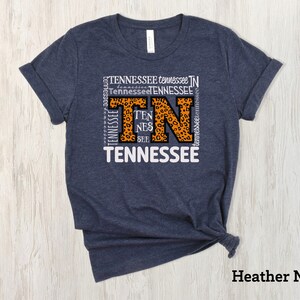 May include: Heather navy blue t-shirt with a white graphic of the letters "TN" in a leopard print design. The letters are surrounded by the word "Tennessee" repeated multiple times.