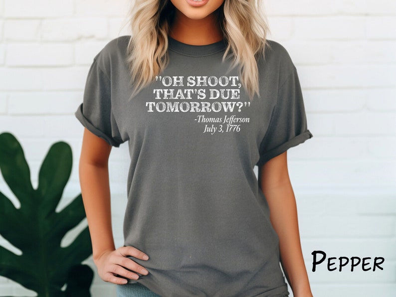 May include: A dark gray t-shirt with a white graphic print that reads "OH SHOOT, THAT'S DUE TOMORROW?" -Thomas Jefferson July 3, 1776
