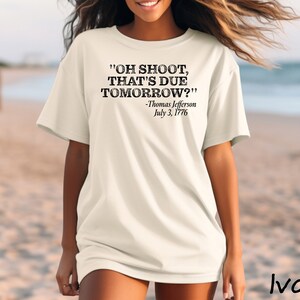 May include: A white t-shirt with a black graphic print that reads "OH SHOOT, THAT'S DUE TOMORROW?" -Thomas Jefferson July 3, 1776. The shirt is worn by a person standing on a beach.