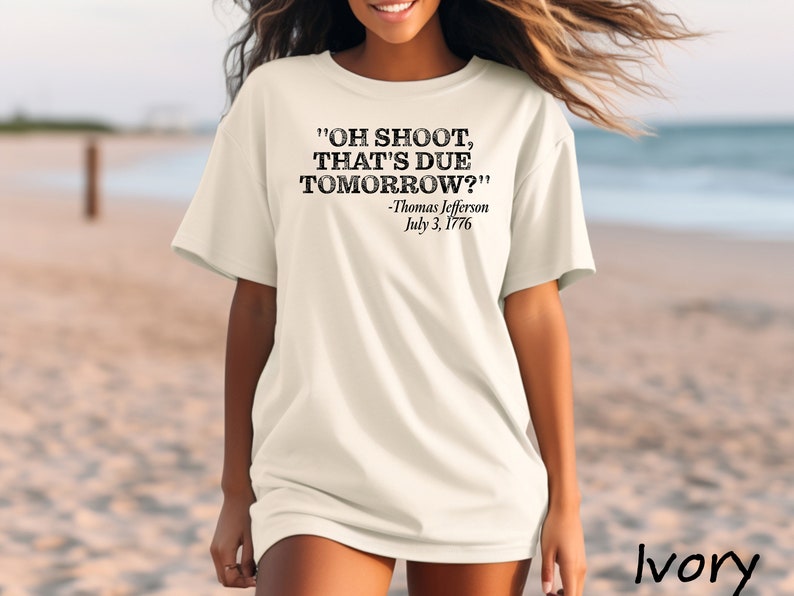 May include: A white t-shirt with a black graphic print that reads "OH SHOOT, THAT'S DUE TOMORROW?" -Thomas Jefferson July 3, 1776. The shirt is worn by a person standing on a beach.