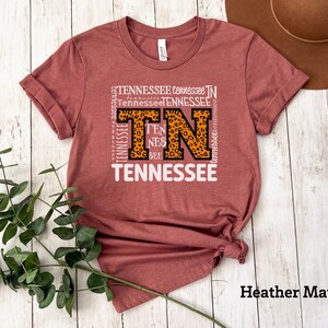 May include: A heather mauve t-shirt with a graphic design featuring the letters "TN" in a leopard print pattern surrounded by the word "Tennessee" repeated in various sizes and orientations.