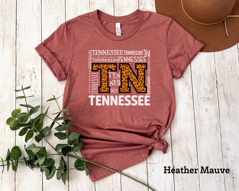 May include: A heather mauve t-shirt with a graphic design featuring the letters "TN" in a leopard print pattern surrounded by the word "Tennessee" repeated in various sizes and orientations.