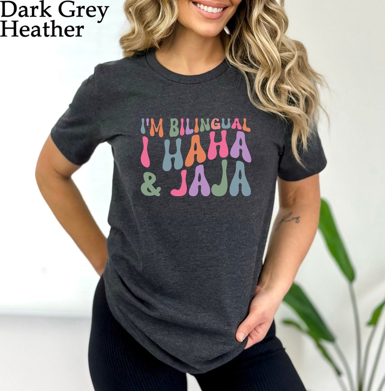 May include: A dark heather grey t-shirt with the text "I'm Bilingual I HAHA & JAJA" in colorful, retro-style lettering.
