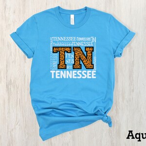 May include: A light blue t-shirt with a white graphic of the letters "TN" in a leopard print design. The letters are surrounded by the word "Tennessee" repeated multiple times. The shirt is folded and displayed on a light wood background. The word "Aqua" is written in black at the bottom of the image.