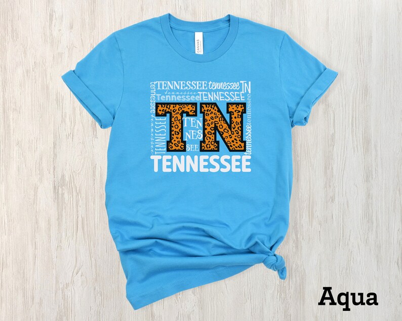May include: A light blue t-shirt with a white graphic of the letters "TN" in a leopard print design. The letters are surrounded by the word "Tennessee" repeated multiple times. The shirt is folded and displayed on a light wood background. The word "Aqua" is written in black at the bottom of the image.