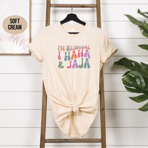 May include: A soft cream colored t-shirt with the text "I'm Bilingual I HAHA &JAJA" in colorful, retro-style lettering.