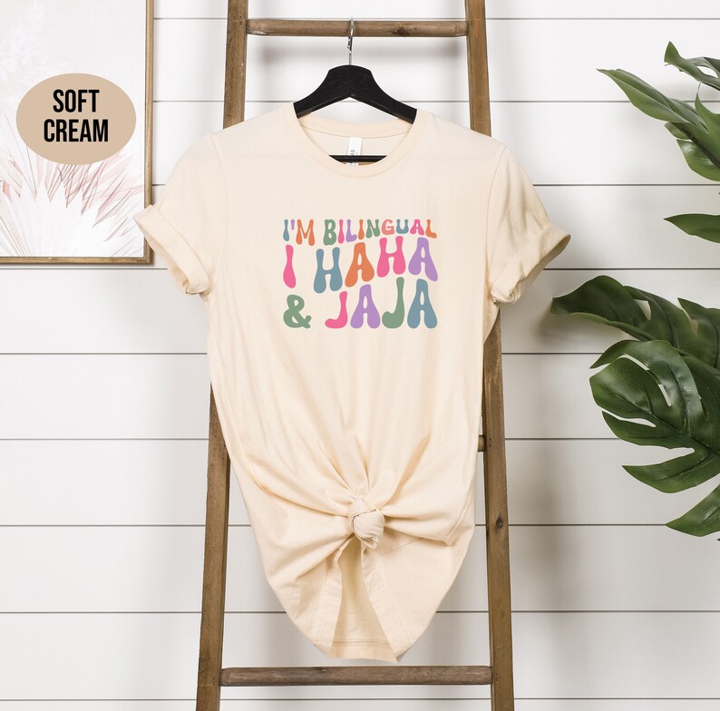 May include: A soft cream colored t-shirt with the text "I'm Bilingual I HAHA &JAJA" in colorful, retro-style lettering.
