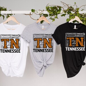 May include: Three t-shirts with a graphic design featuring the letters "TN" in a large font with a leopard print pattern. The letters are surrounded by the word "Tennessee" repeated multiple times in a smaller font. The t-shirts are white, grey, and black.